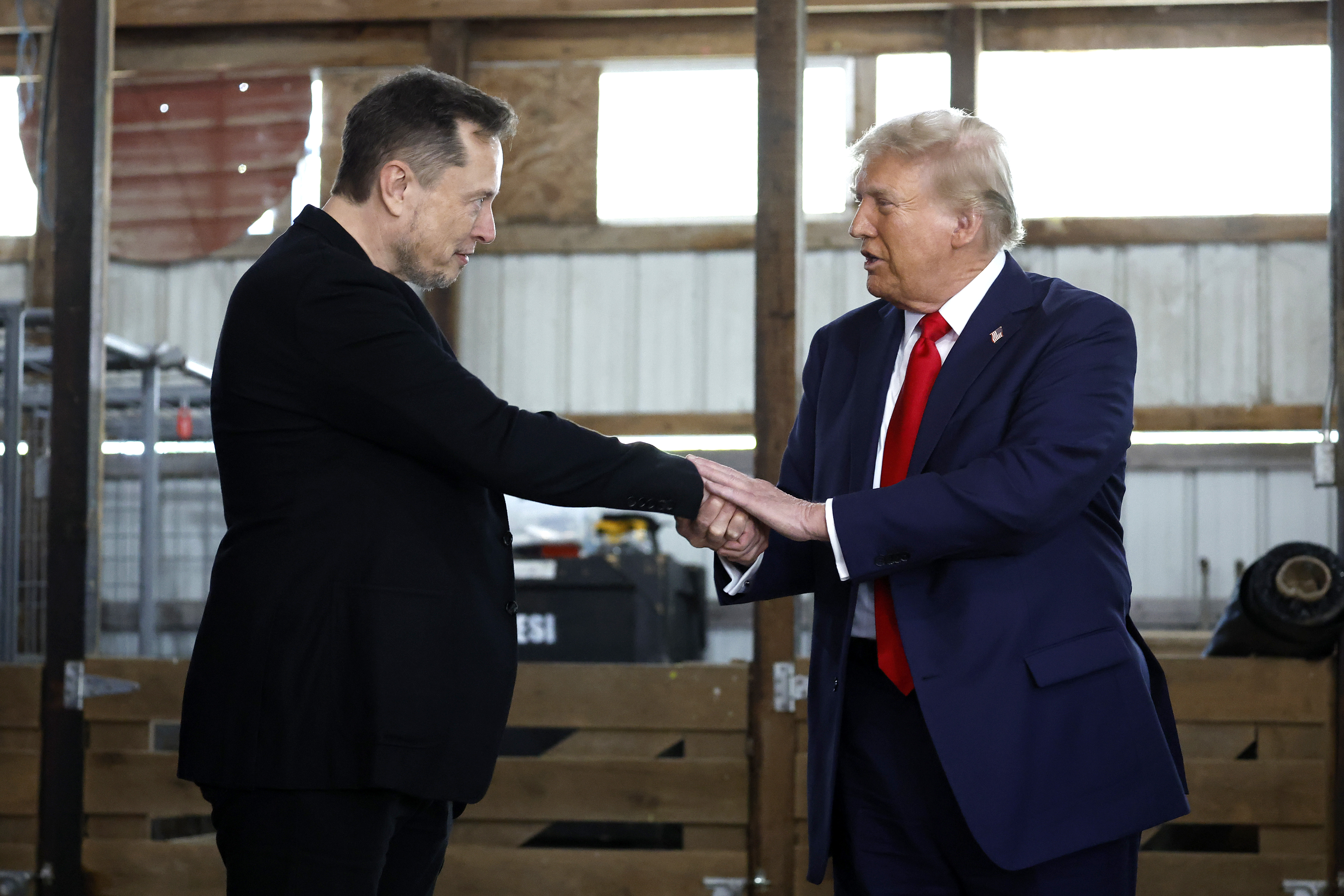 Elon Musk e Donald Trump, ambos de terno, trocam aperto de mãos enquanto se olham.