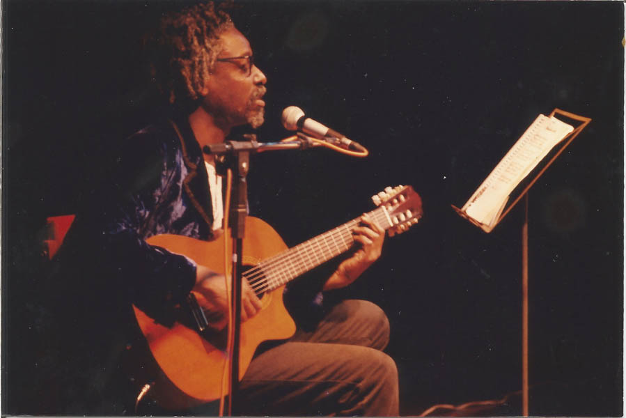 Fotografia colorida. Itamar Assumpção canta ao microfone e toca violão sentado durante show. A sua frente há uma estante de partitura.