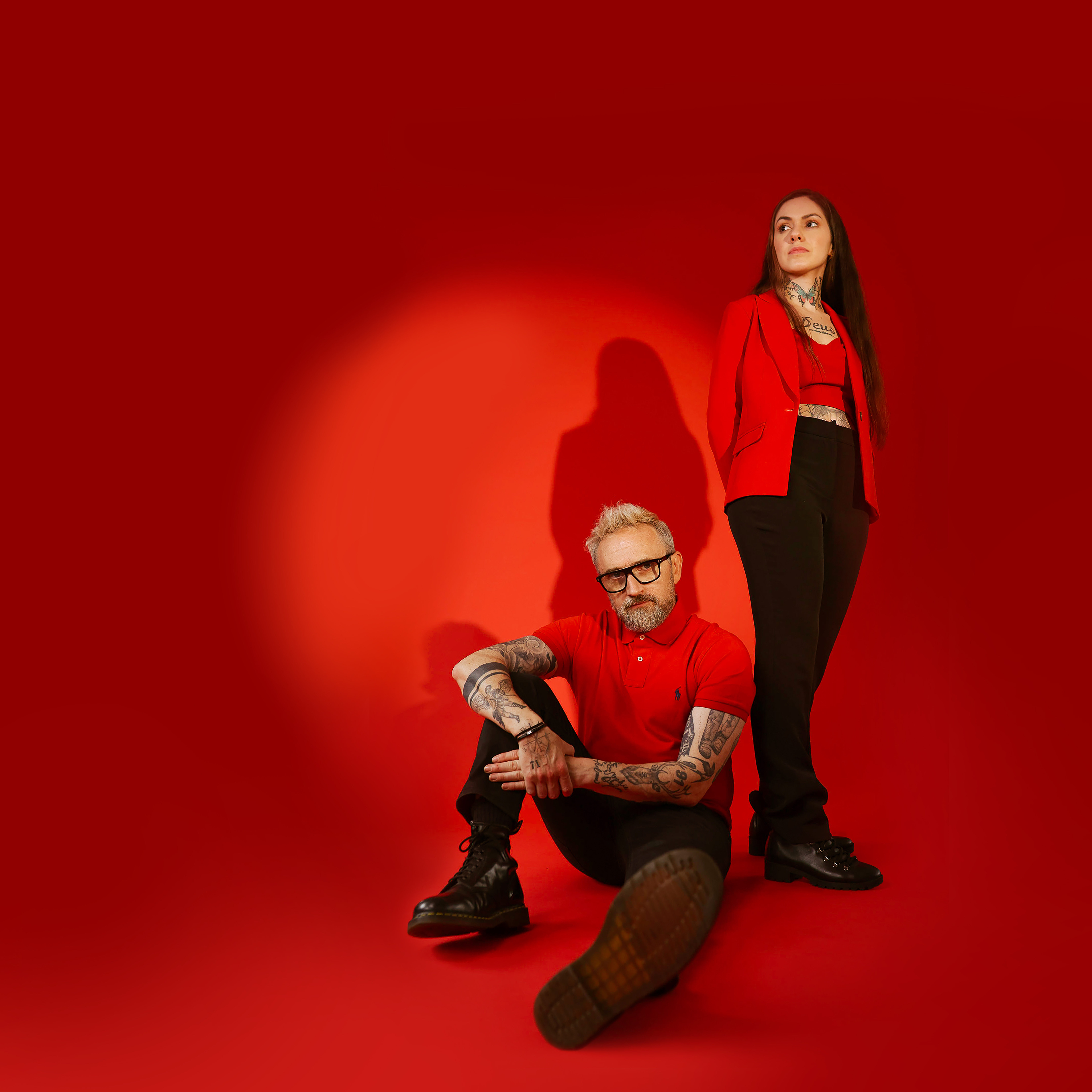 Noel Hogan sentado com camiseta vermelha. Ao lado dele, em pé, está Mell Peck, tambpem com camiseta vermelha. Foto em estúdio com fundo vermelho.